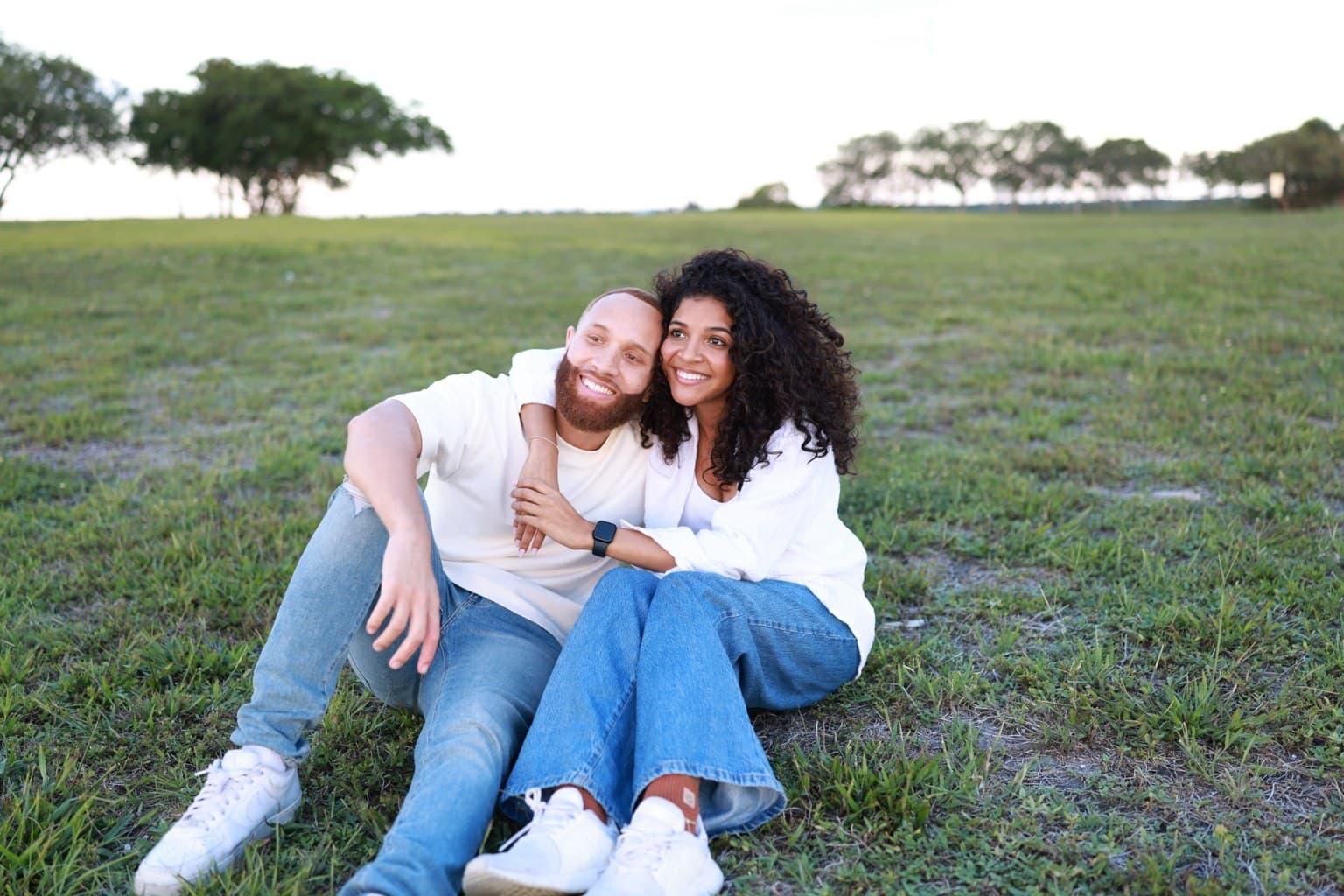 Ray and Alexia seated close together in a grassy field at dusk, Alexia's arms wrapped around Ray's shoulders